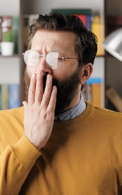 男はあくびをします。オフィスやアパートの部屋で眼鏡をかけた疲れた悲しいあごひげを生やした男は、あくびをして、手で口を覆います。クローズアップビュー