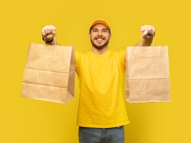 Man in yellow cap, tshirt giving paper packets isolated. Male employee courier hold paper packets with food