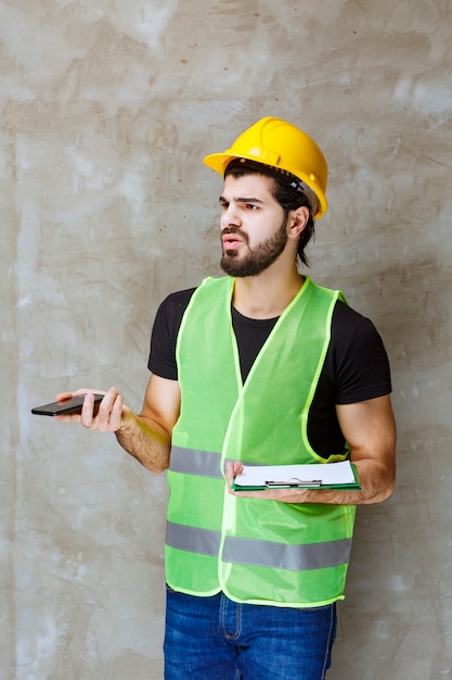 Man in yellow helmet and gear holding a project plan and talking to the phone