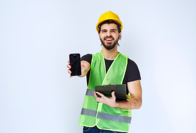 Uomo in casco giallo che tiene una cartella di progetto e uno smartphone.