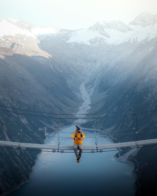 Photo man in yellow jacket sitting on a suspension bridge over a lake in olpererhutte austria