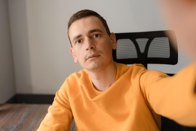 A man in a yellow sweater is taking a selfie while sitting at a desk at home