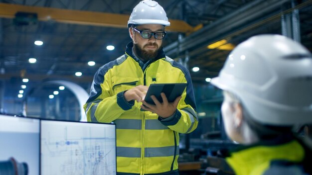 Photo a man in a yellow and white hard hat is standing in front of a blueprint and a man in a yellow vest is holding a tablet.