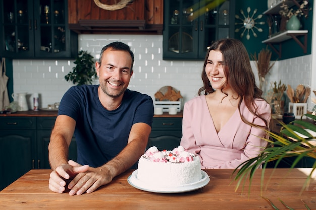 Man and young woman sit in home kitchen with cake on table.