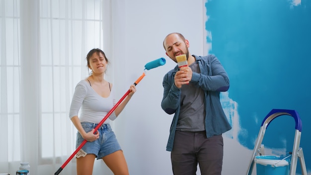 Man zingt op schilderborstel terwijl hij appartement opknapt met vrolijke vrouw. appartement herinrichting en woningbouw tijdens renovatie en verbetering. reparatie en decoreren.