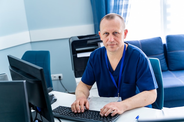 Man zit aan bureau. Dokter in scrubs op kantoor in de buurt van computer. Kantoor in het ziekenhuis.