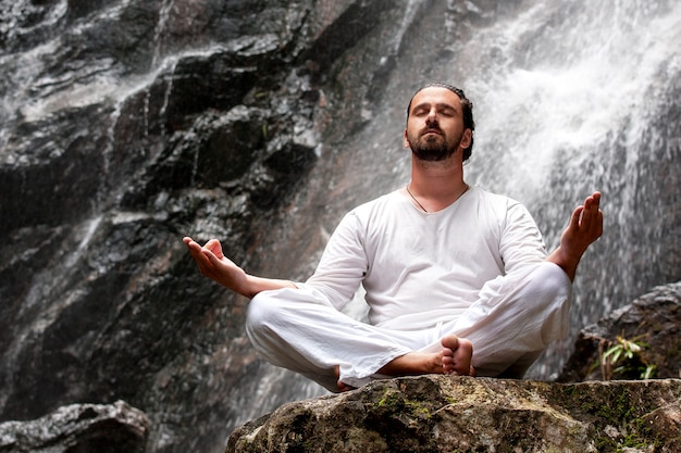 Man zit in meditatie yoga op rots bij waterval in tropisch regenwoud