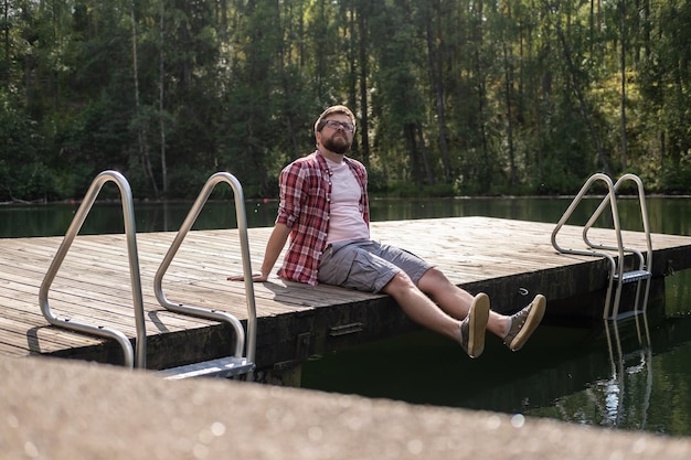 man zit op de rand van een houten pier en kijkt bedachtzaam op tegen een kalm bosmeer