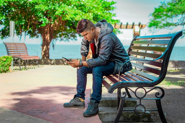 Man zit op een bankje zijn telefoon te checken, jonge man kijkt op zijn mobieltje zittend op een bankje