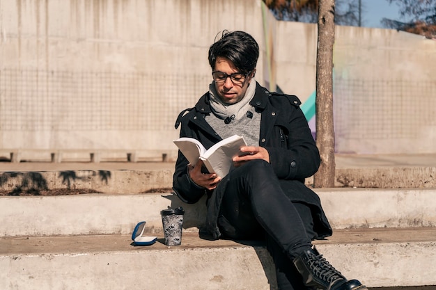 Man zit op een betonnen bankje in een openbaar park, leest een boek en drinkt een warm drankje in de zon.