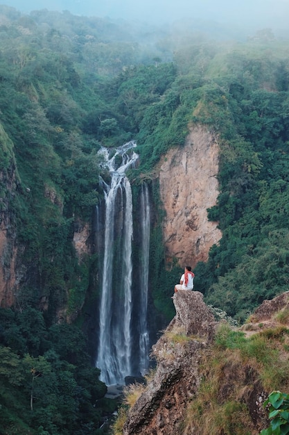 Man zit op een klif tegen een waterval in het bos.