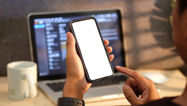 Man zit voor computerlaptop en houdt slimme telefoon met wit scherm vast