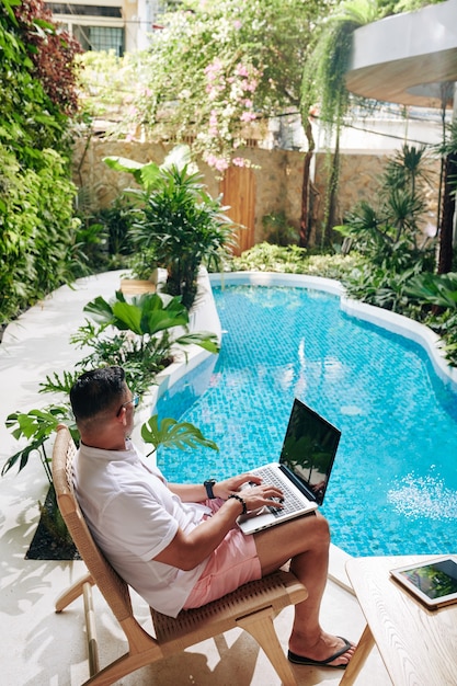 Man zittend in een stoel naast het zwembad in zijn achtertuin en bezig met laptop, freelancen en werken op afstand concept