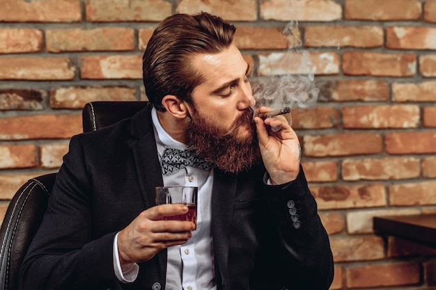 man zittend op een stoel en met een glas whisky in zijn hand en het roken van een bruine sigaar