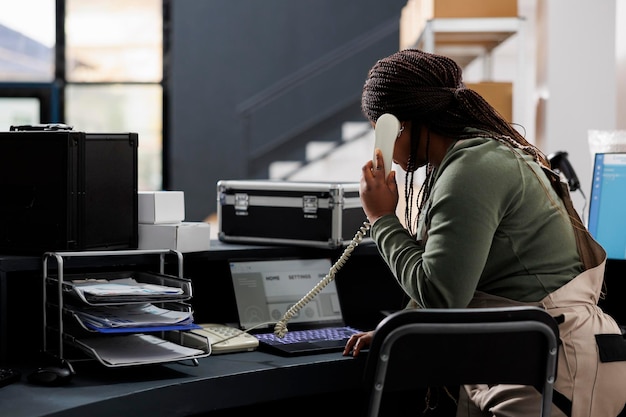 Manager bereidt bestelling voor en bespreekt verzenddetalis met externe klant op vaste telefoon in magazijn. Afro-Amerikaanse werknemer analyseert goederenchecklist op laptopcomputer in opslagruimte