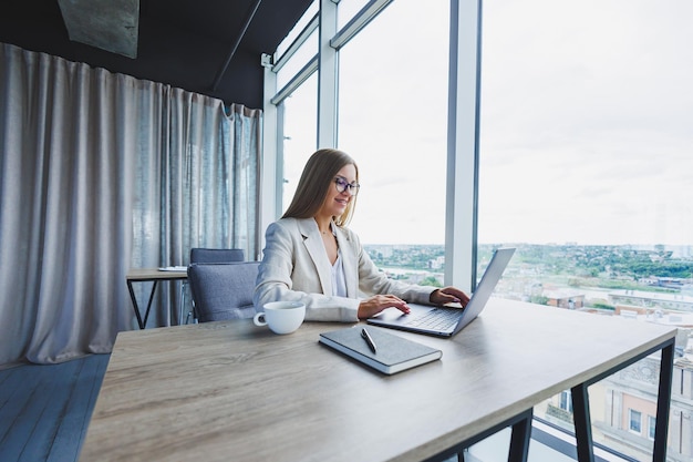 Manager focused woman in casual wear and eyeglasses typing on netbook while working on new project sitting at table in modern workspace