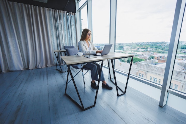 Manager focused woman in casual wear and eyeglasses typing on netbook while working on new project sitting at table in modern workspace