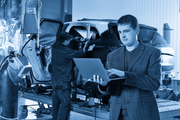 Manager holding laptop while standing near car in auto repair shop with mechanic repairing car on