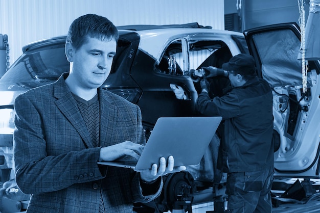 写真 車を修理する整備士がいる自動車修理工場で車の近くに立ちながらラップトップを持つマネージャー