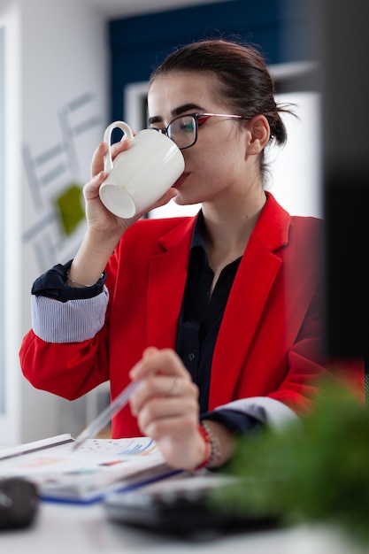 白いカップから飲んでスタートアップデスクで赤いジャケットのマネージャー。オフィスでコーヒーやお茶を飲んでいる眼鏡をかけている従業員。クリップボードのチャートを読むのを一時停止している実業家。