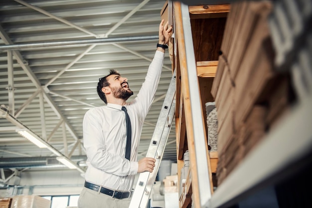 A manager standing on ladders at printing shop and trying to reach box with materials