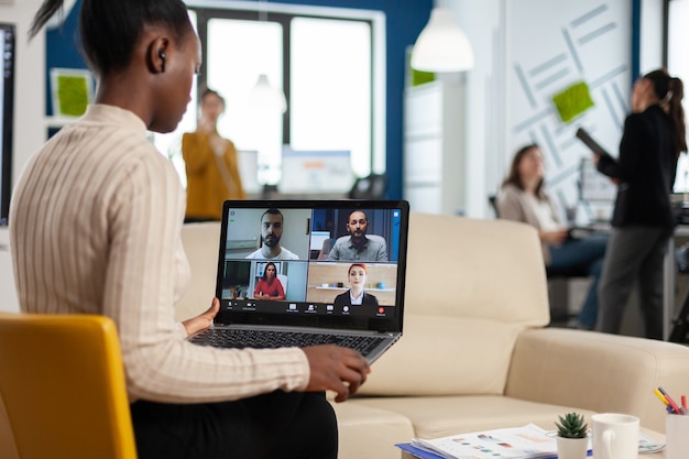 Photo manager woman discussing with remote colleagues using video call holding laptop sitting on couch in business modern office