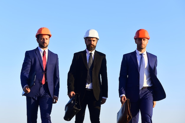 Managers hold folder and briefcases Leaders with beard and determined faces walk in team Building and business concept Board of architects wear suits ties and hardhats on blue sky background