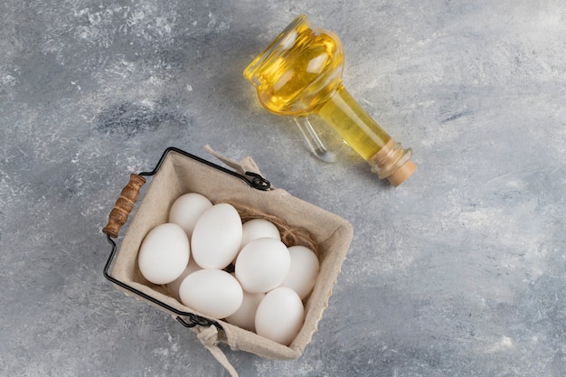 Mand vol verse witte kippeneieren met een glazen fles olie op een marmer.