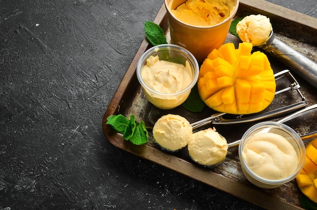 Mango ice cream with mint and fresh mango. Ice cream spoon. On a black stone background, top view.