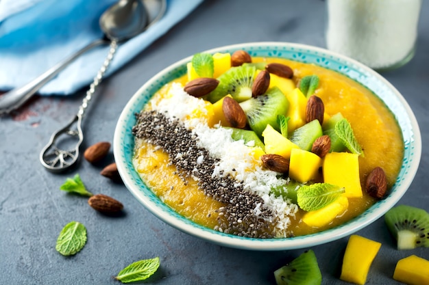 Mango smoothie bowl with pieces of kiwi, mango, coconut shavings and chia seeds on a stone