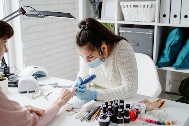 Manicure master in mask and gloves using an electric machine to remove the nail polish