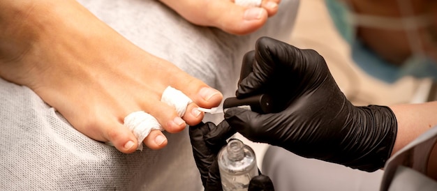 Manicure master painting nails on a female foot with transparent nail polish in a beauty salon close up