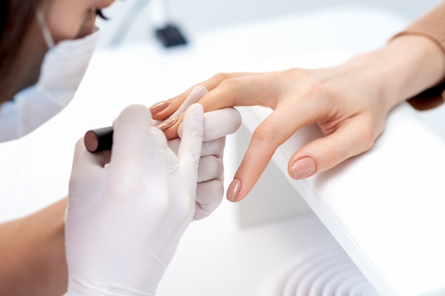 Manicure master in protective gloves applying beige nail polish on female nails in beauty salon