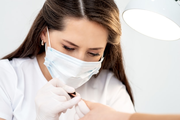 Manicure master with protective mask in rubber gloves applying beige nail polish on female nails in beauty salon