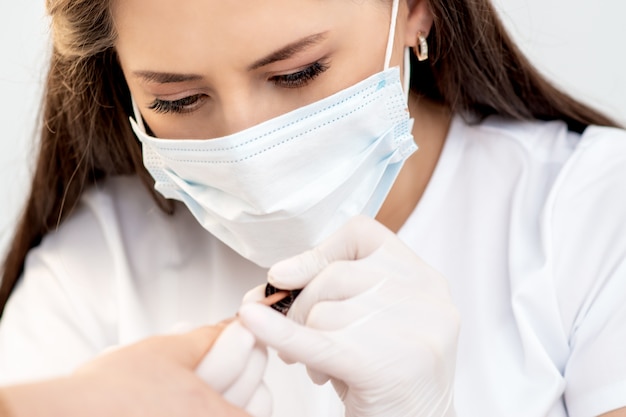 Manicure master with protective mask in rubber gloves applying beige nail polish on female nails in beauty salon