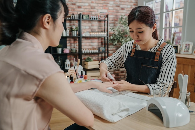 Manicure in process concept. two asian chinese woman customer and manicurist sitting and smiling at spa salon. Beautician trimming cuticles by clipper of female client in modern beauty workshop.