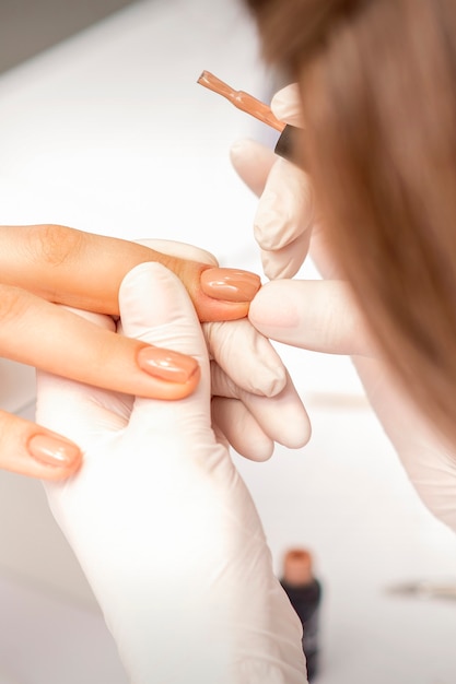 Manicurist applying beige nail polish on fingernails of a female client in a nail salon