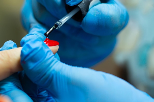 A manicurist in blue gloves, covering the nails with a long-lasting red gel polish.