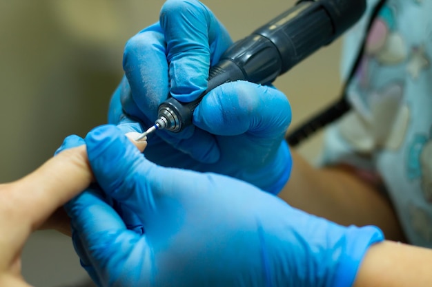 A manicurist in blue gloves makes a manicure with a device. Cuticle treatment