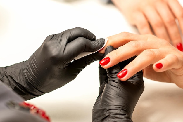 The manicurist finishes the procedure for red nail polishing and cleaning with a cotton napkin pad swab in a beauty salon close up