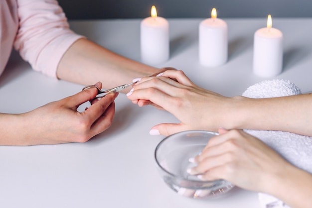 Manicurist hands uses metal cosmetic nail scissors for cutting cuticle from female client's nails. Woman hand receiving manicure procedure with nail care tool.