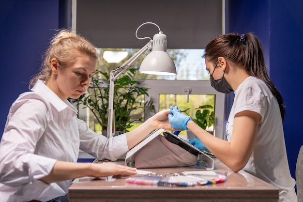 The manicurist is concentrating on the hands of a young woman who has come to the procedure. On the manicure table are samples of varnish with flowers that are available