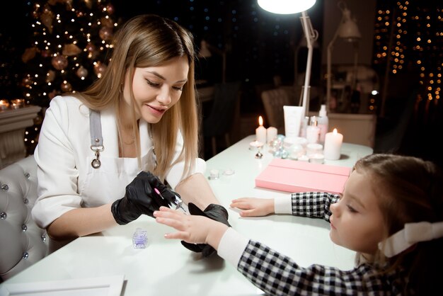 A manicurist in protective gloves paints a little girl's nails in a beauty salon against the background of the Christmas tree.