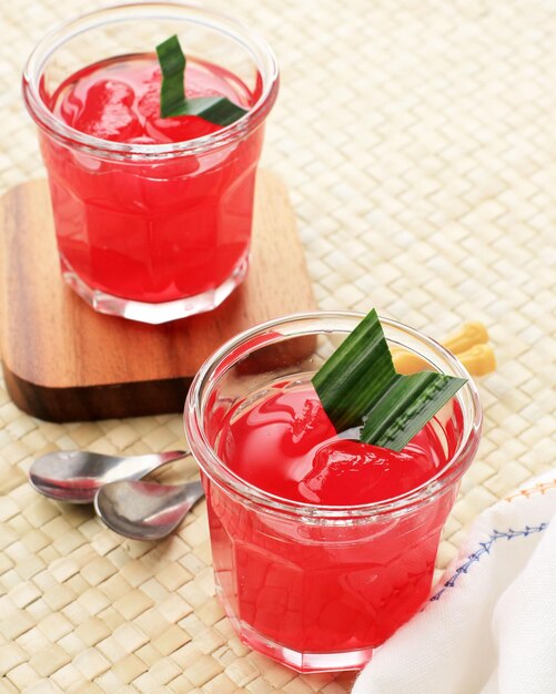 Manisan KolangKaling Preserved Sugar Palm Fruit with Red Color Served in a Glass with Wooden Coaster and Pandan Leaves as Garnish