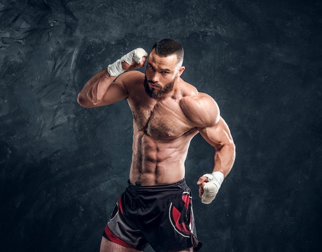 Manly bearded man with beautiful muscular torso is posing for photographer on the dark background.