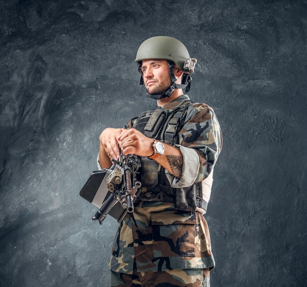 Manly handsome military man in helmet with tattoo on his hand is standing at dark background.