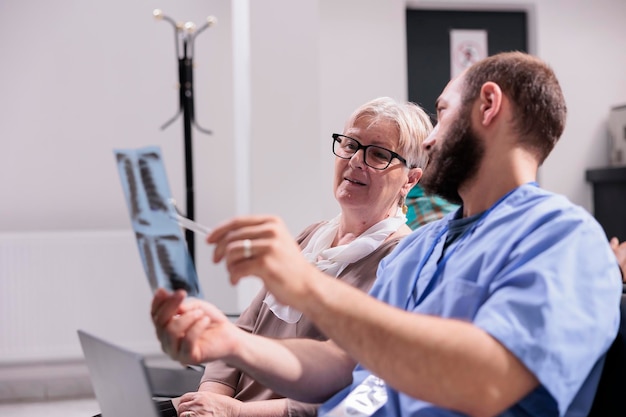 Mannelijk assistent-hospitaalpersoneel dat röntgenfoto's toont aan oudere patiënt, in de lobby van de ziekenhuisreceptie. Man verpleegster die de resultaten van de radiografie scan uitlegt aan vrouw met ziekte, praten over behandeling en diagnose.