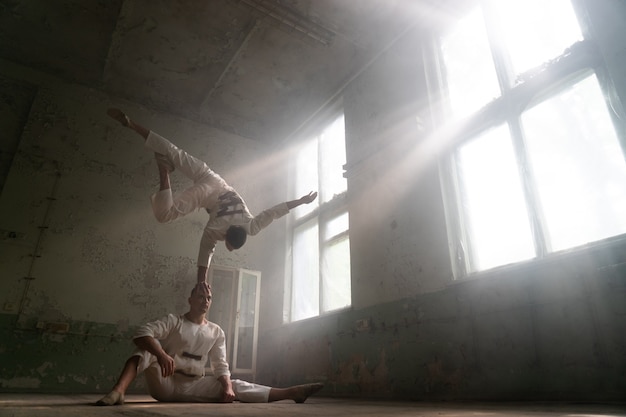 Mannelijk duo maakt acrobatische trucs in kostuum van krankzinnige mensen in verlaten kamer met zonnestralen uit ramen.