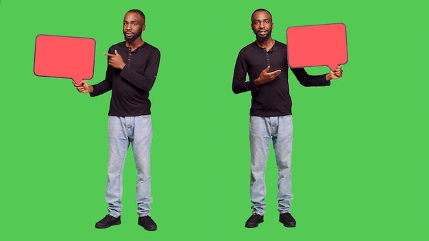 Mannelijk model met rode lege tekstballon op camera, presentatie maken met lege copyspace kartonnen pictogram. Jonge volwassene die modelaanplakbord over groen het schermachtergrond toont.