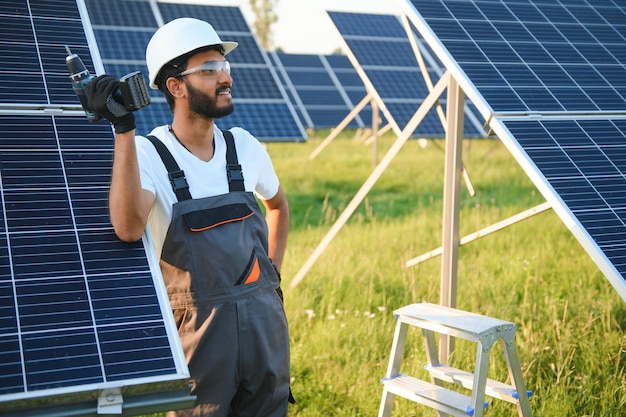 Mannelijke Arabische ingenieur in helm en bruine overalls die de weerstand in zonnepanelen buiten controleert Indiase man die op het station werkt